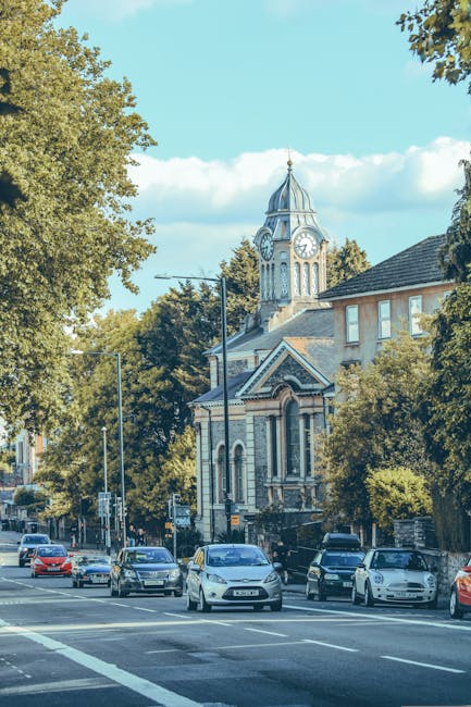 A street view in East Barnet showing multiple parked cars along the pavement, with a church featuring a tall clock tower and a pointed spire visible in the background. The scene is framed by leafy gre